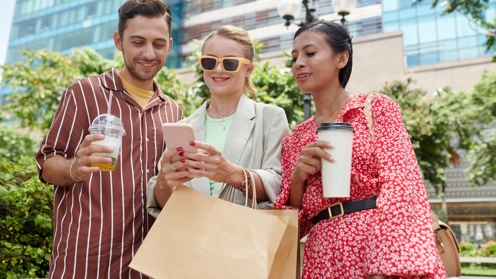three friends outside looking at a mobile phone while they enjoy sodas at the mall and carry shopping bags